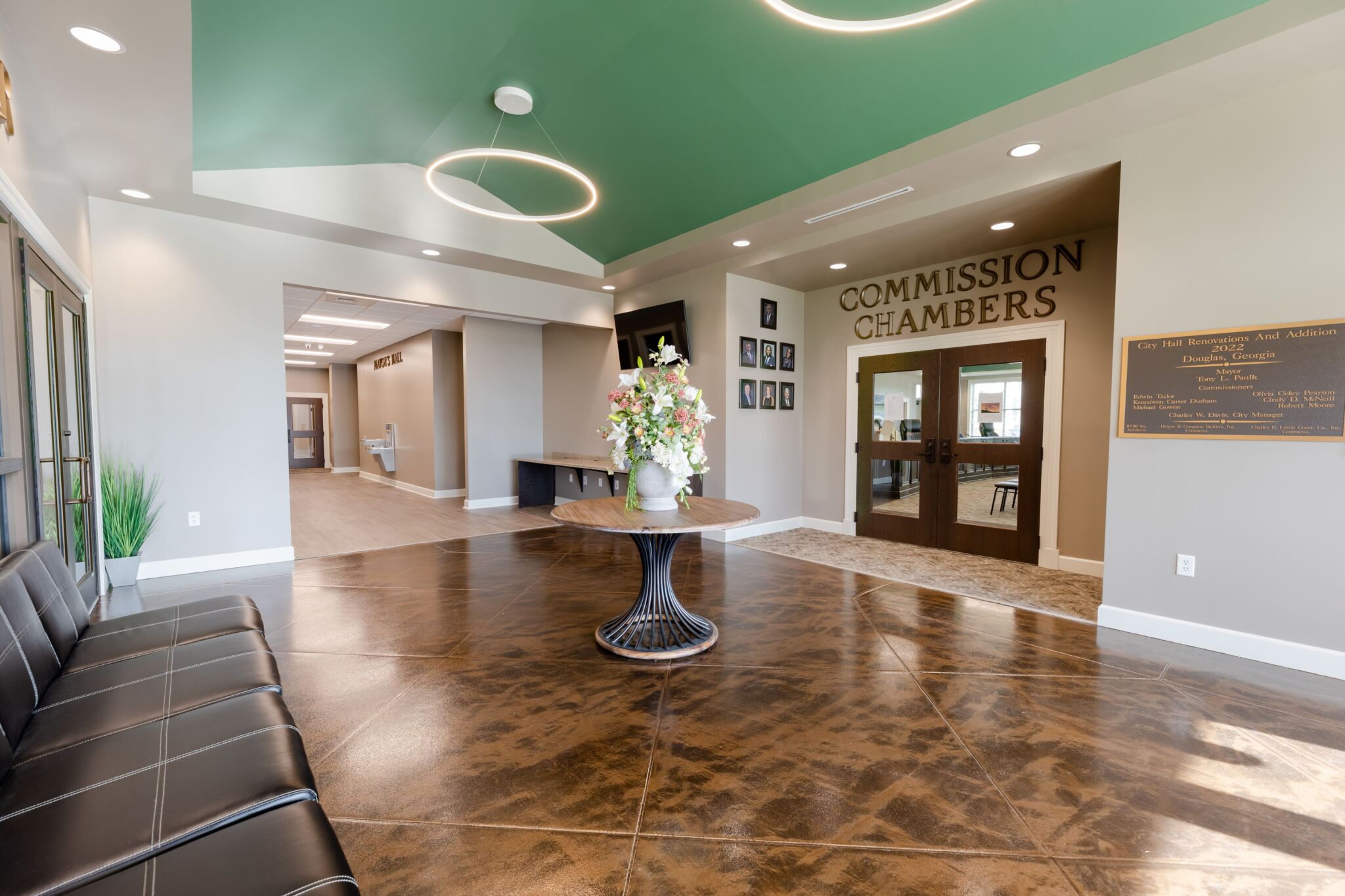 Spacious lobby with brown polished floors, round table with flowers, black cushioned seating, and double doors labeled Commission Chambers under a mint green ceiling with circular lights.