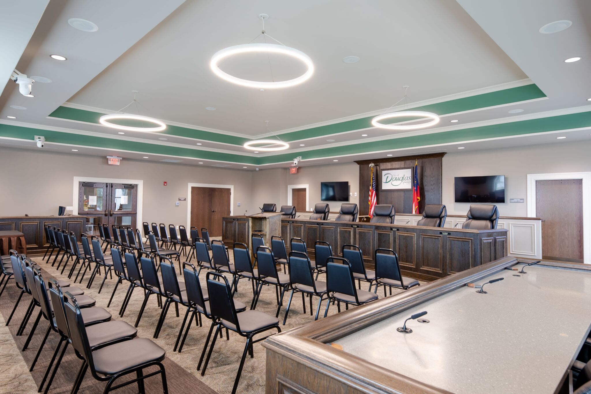 A modern, well-lit council chamber with rows of empty chairs facing a podium and desks. Two flags and a Dawsonville sign behind.