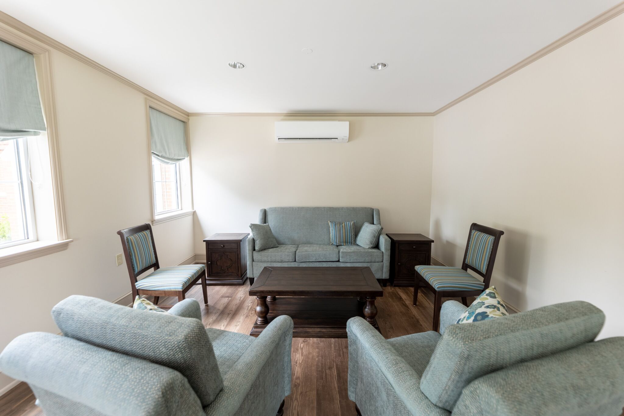 Small living room with light blue sofas, armchairs, dark wood coffee table, side tables, wood floor, blue-shaded windows, AC on wall.