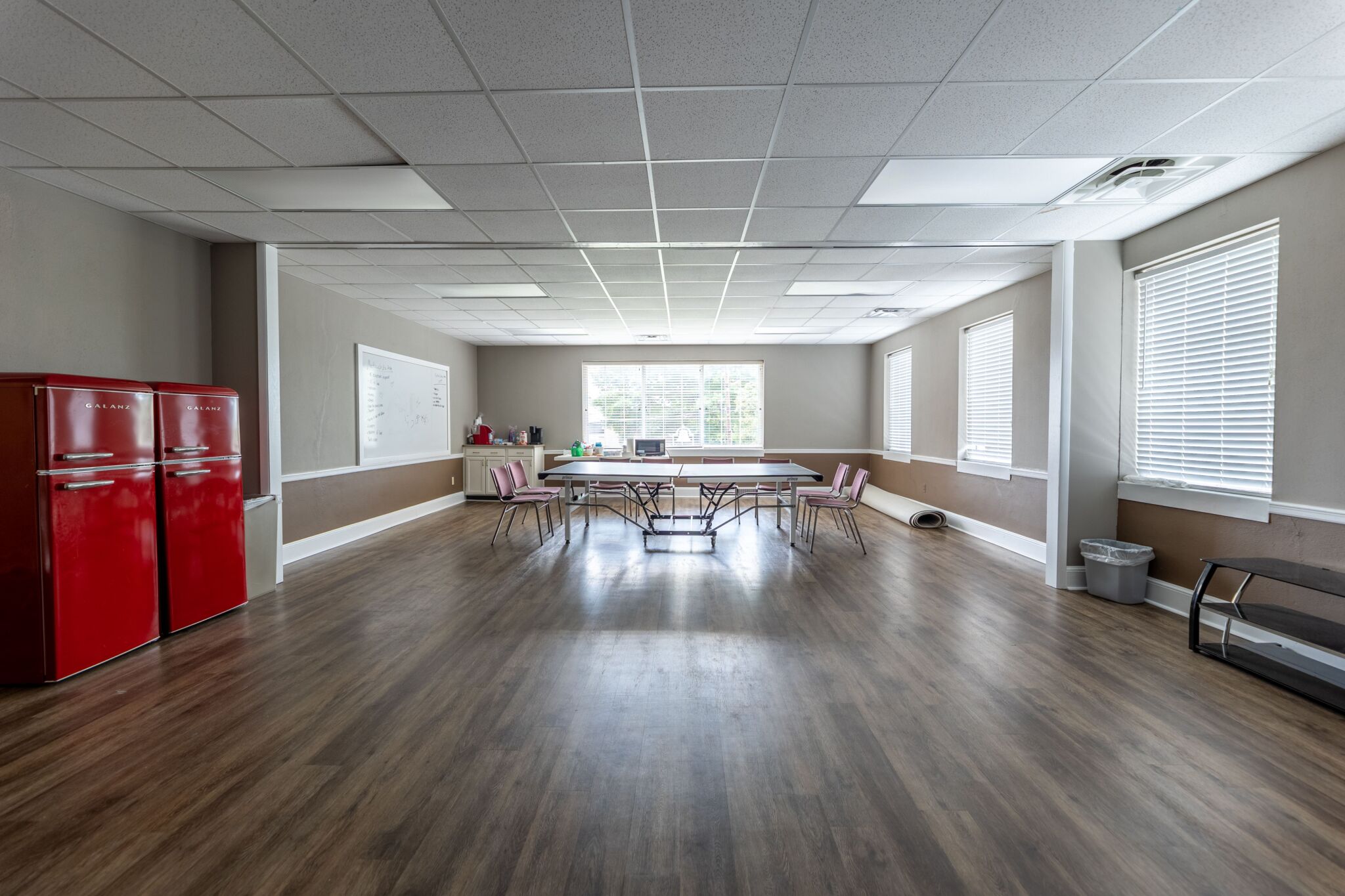 Spacious room with wood flooring, two red refrigerators on the left, big windows, pink chairs around a table, and supplies by a whiteboard.