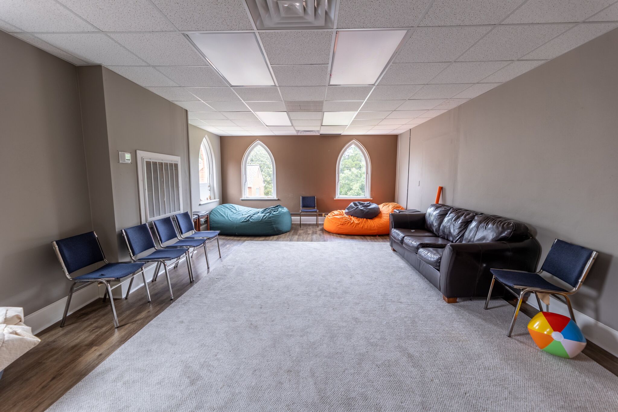 A spacious room with gray carpet, blue and orange bean bags, a black leather couch, blue chairs by the wall, arched windows, and a colorful beach ball on the floor.