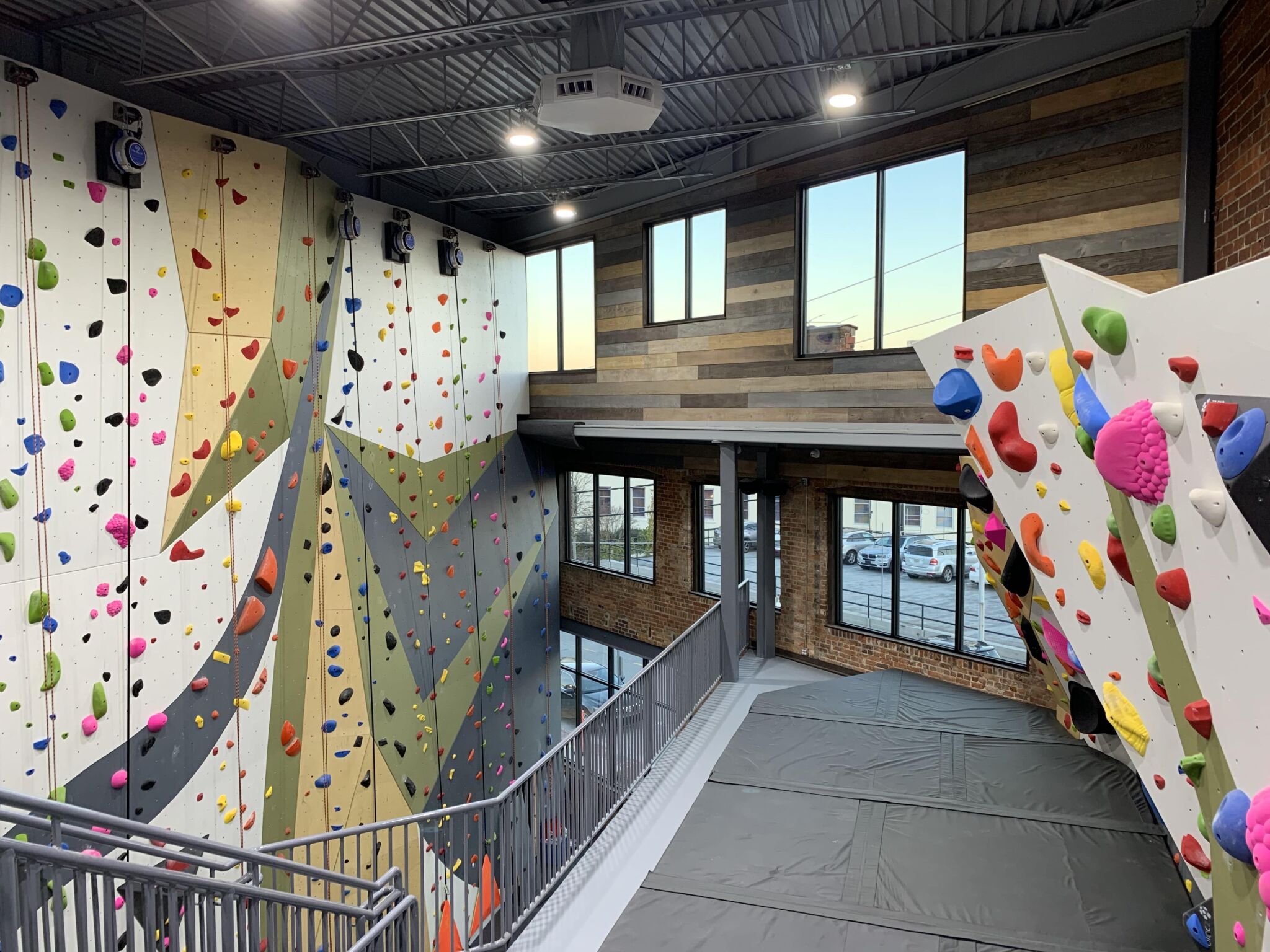 An indoor rock climbing gym with tall colorful climbing walls, padded floors, big windows, and a mezzanine under industrial lights.