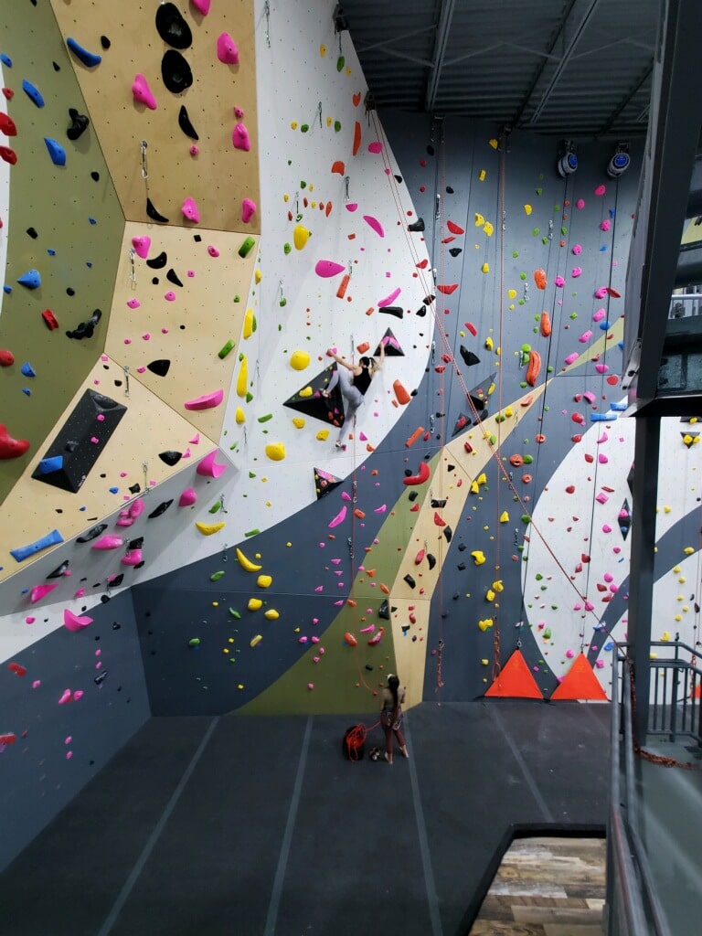 Indoor rock climbing gym with tall walls, colorful holds, a climber in the center, a belayer below, and bright high ceilings.