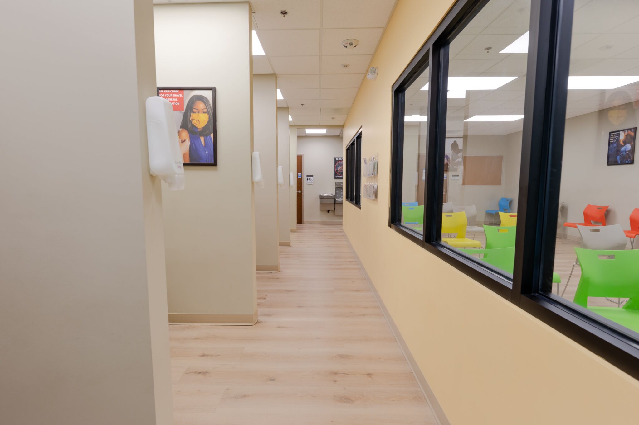 A medical office hallway with light wood floors, beige walls, and windows showing a waiting area with colorful chairs and decor.