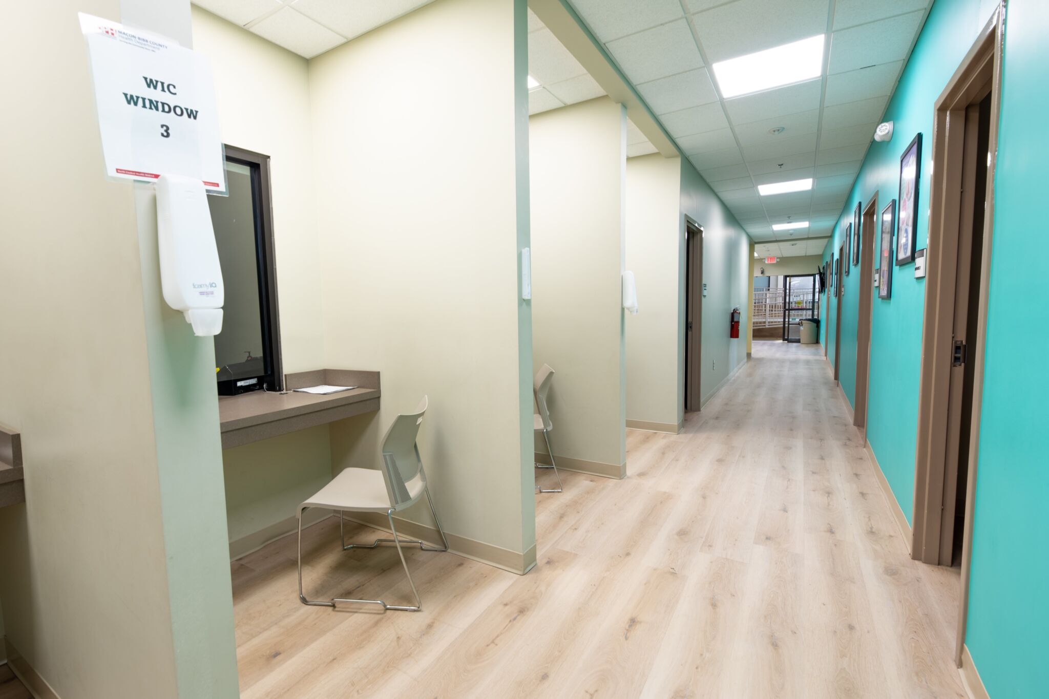 Brightly lit clinic hallway with teal walls, wood floors, WIC service window and chairs on left, doors along hall toward open area.
