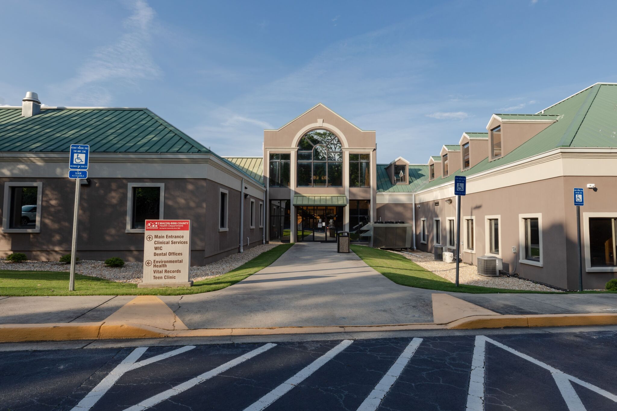 Modern medical clinic with green roofs, tan walls, large windows, accessible parking, and entrance sign with directional info.