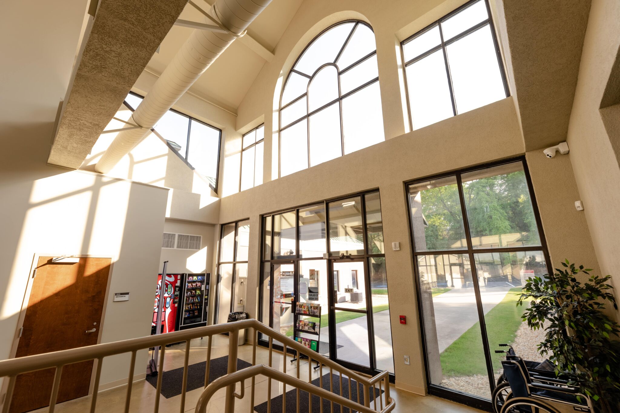 Spacious sunlit lobby with large arched windows, vending machines, indoor plants, and outdoor area visible through glass doors.