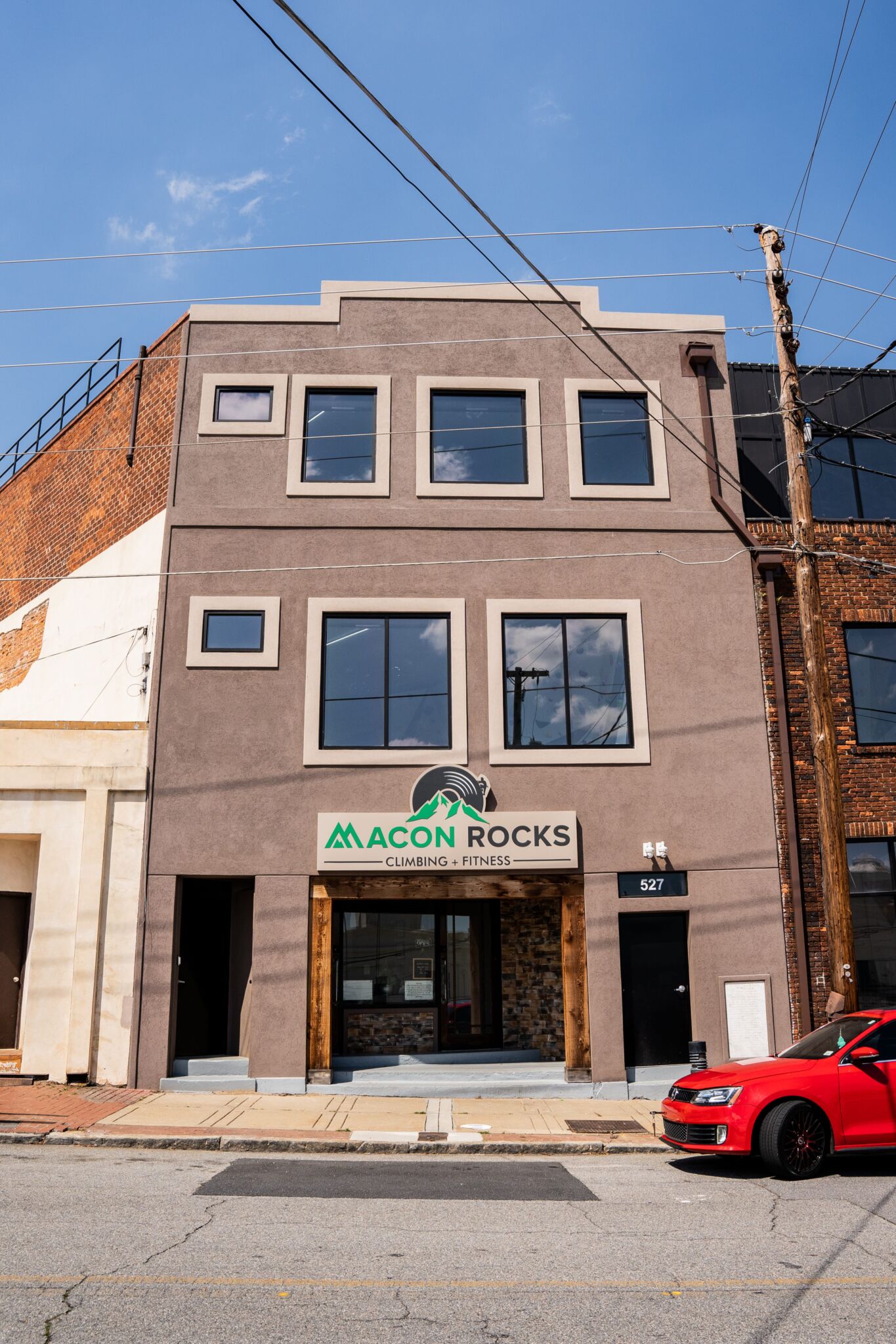 A three-story building with a brown facade houses Macon Rocks Climbing + Fitness. Double glass doors, red car in front, clear sky.