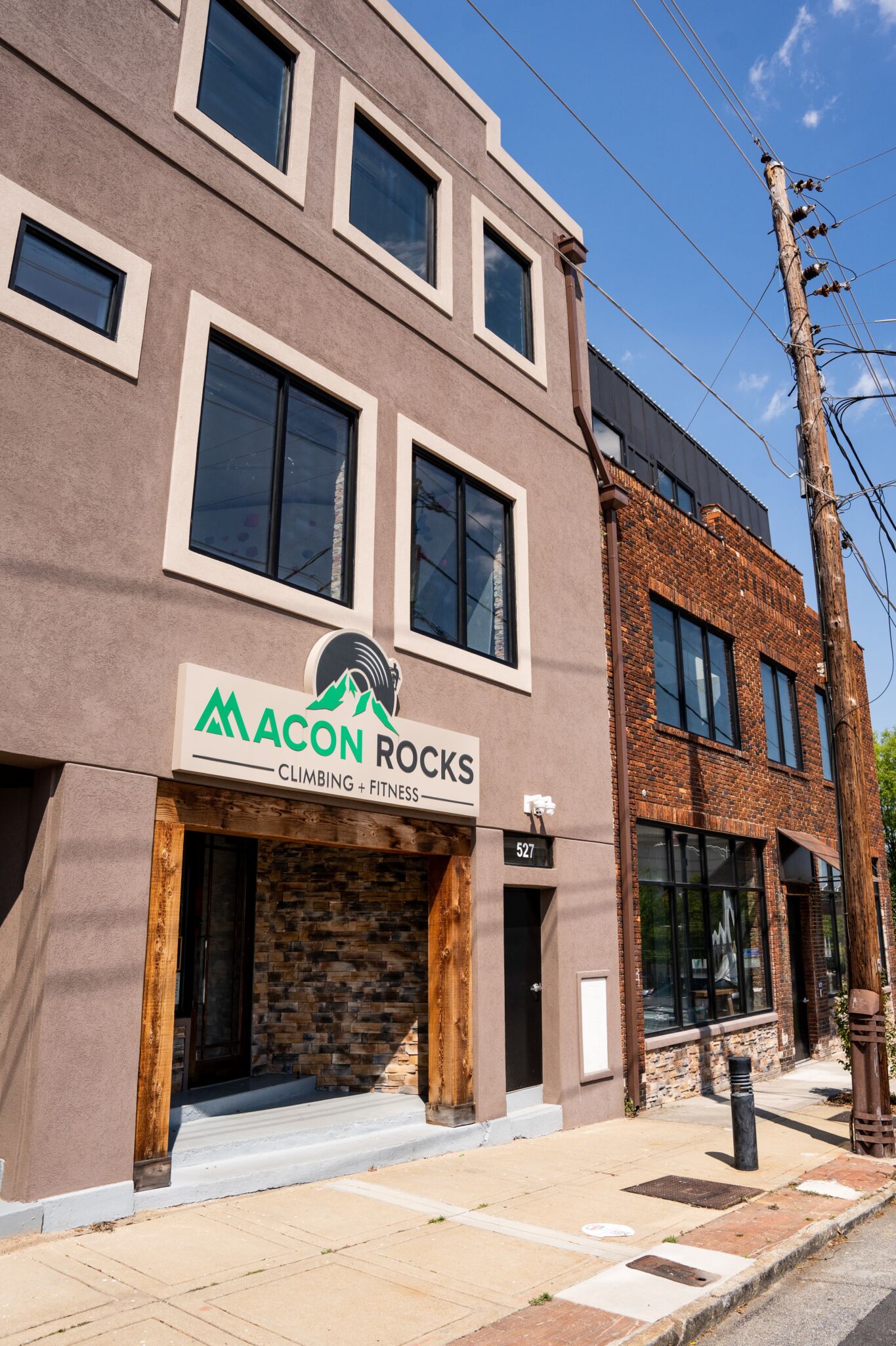 A modern building with large windows and a sign reading Macon Rocks Climbing + Fitness above the entrance, on a sunny street with utility poles and a sidewalk.