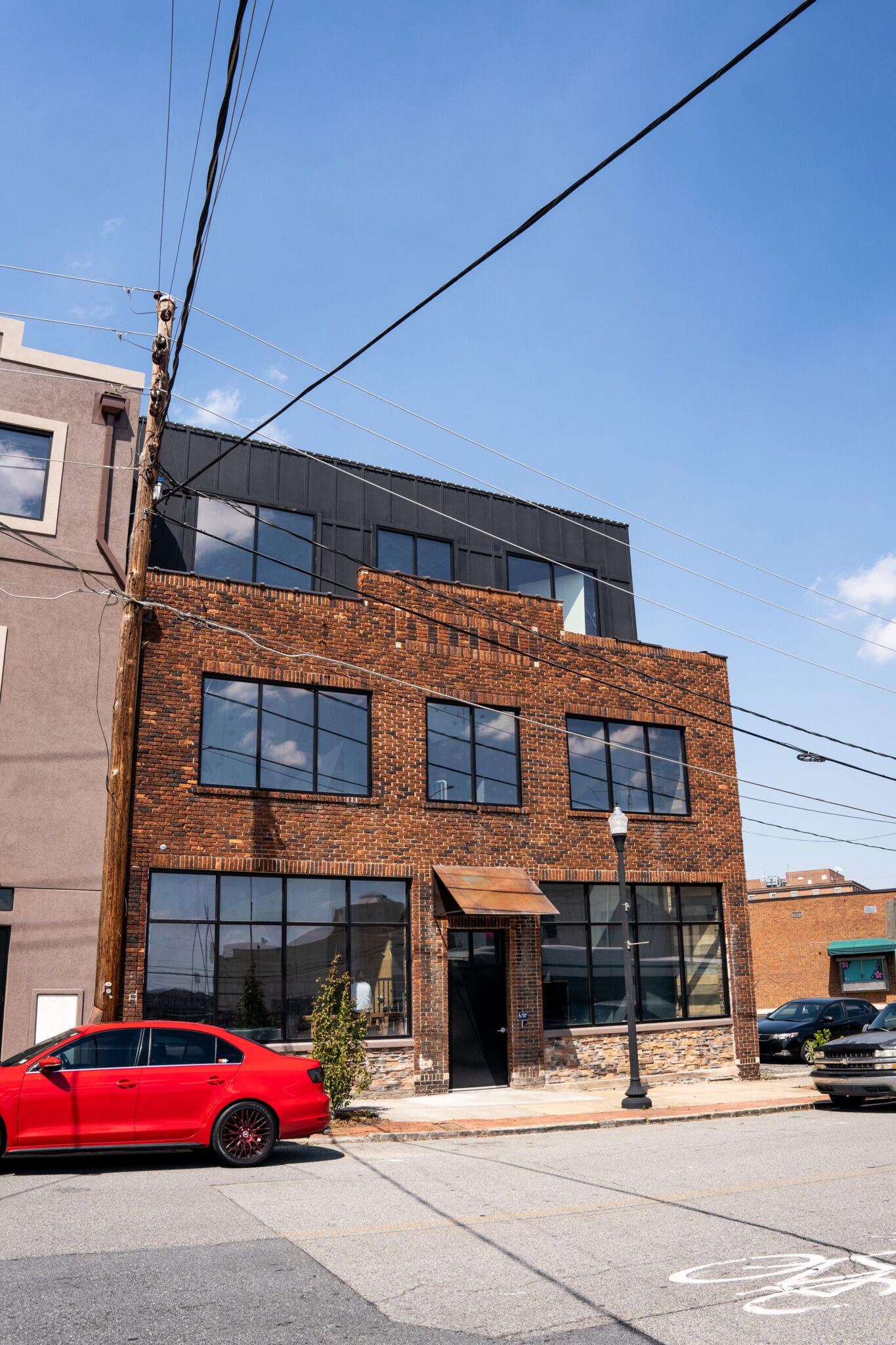 Three-story brick building with large windows and metal awning beside a gray building, red car parked in front, power lines above.