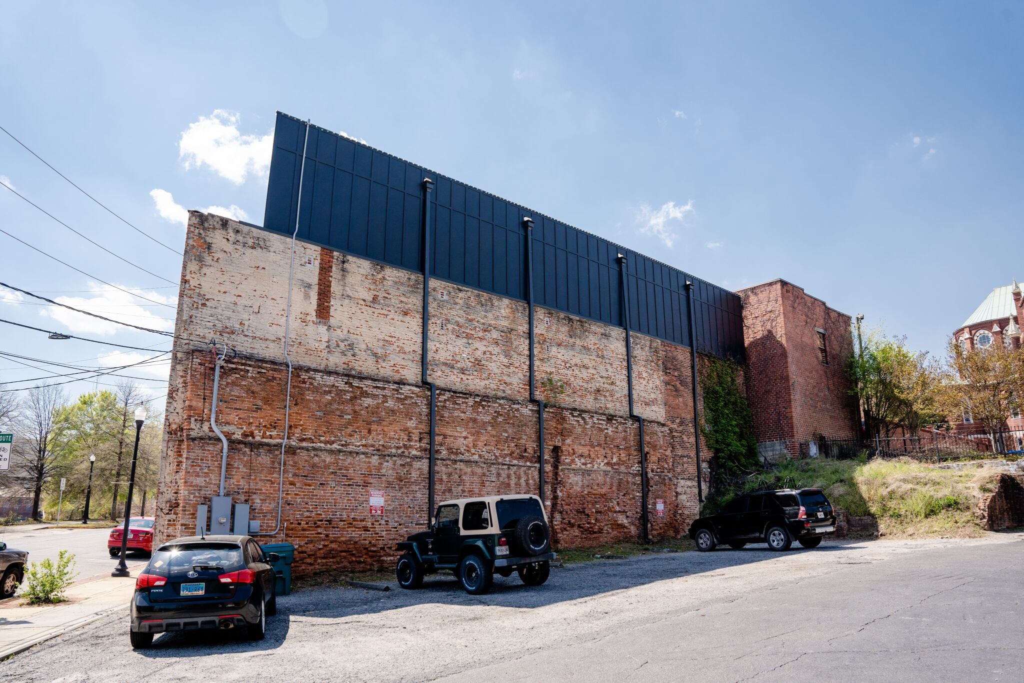 Four vehicles are parked by a tall, worn brick building with a black metal top. Greenery and a hill are behind it under blue sky.