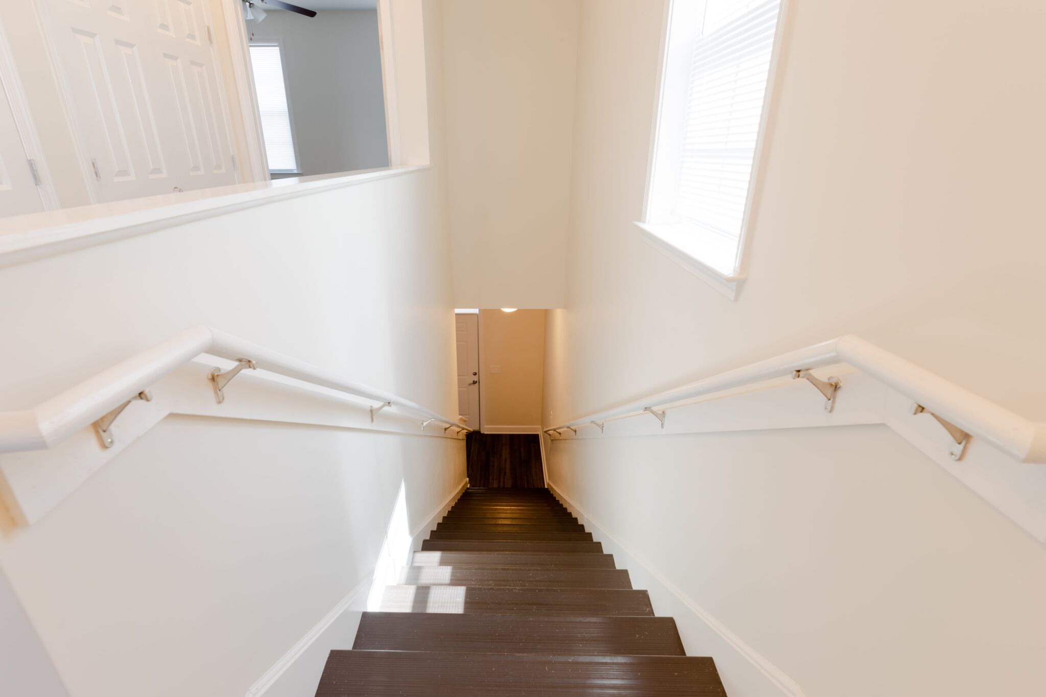 View from the top of a staircase with dark wooden steps and white walls, lit by natural light; a closed door is at the bottom.