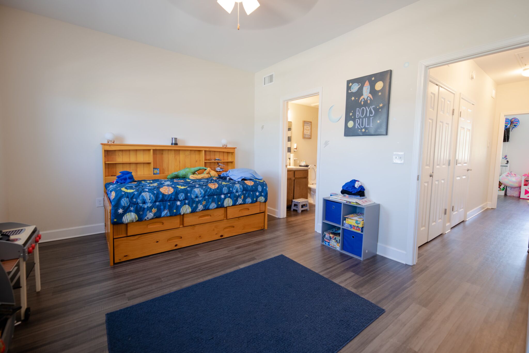A tidy children's bedroom with a single bed featuring a space-themed bedspread, blue rug, toy shelves, and a Kids Rule sign. An open doorway leads to a bathroom and hallway.