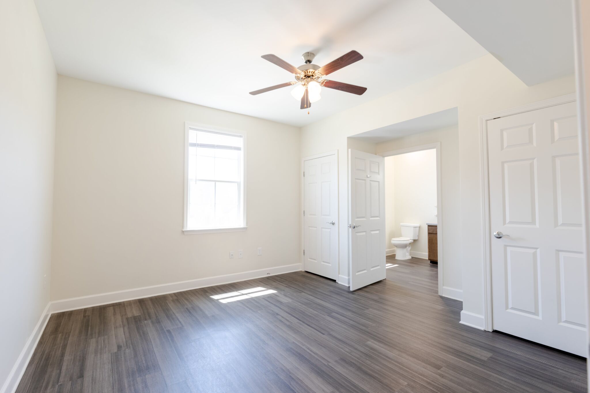 Empty, bright bedroom with light walls, natural light from a window, wooden floor, ceiling fan, and two doors—one to a bathroom.