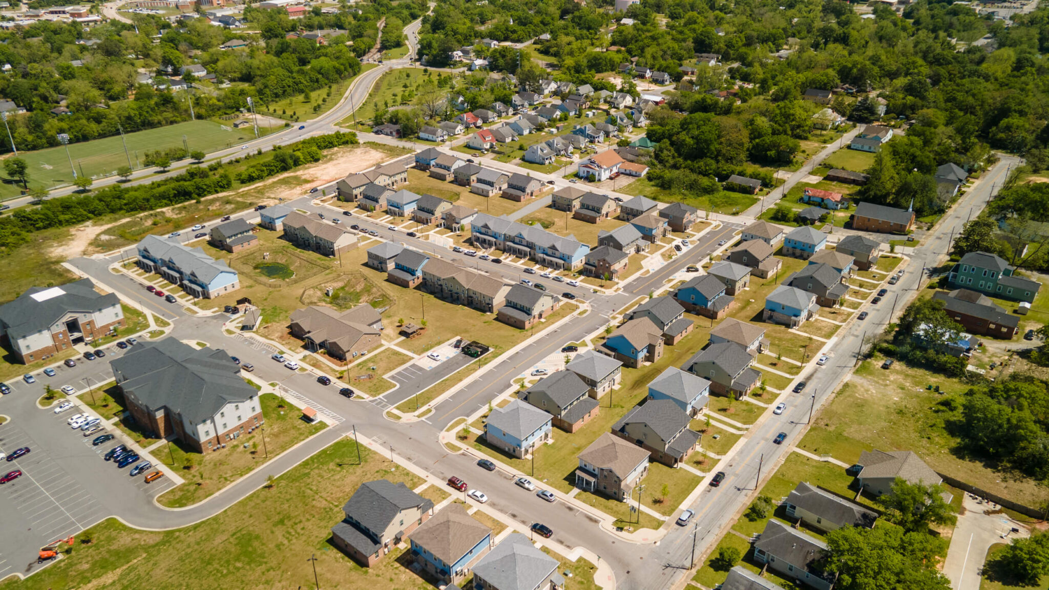 Aerial view of a suburban neighborhood with rows of houses, streets, parking lots, green spaces, and surrounding trees.