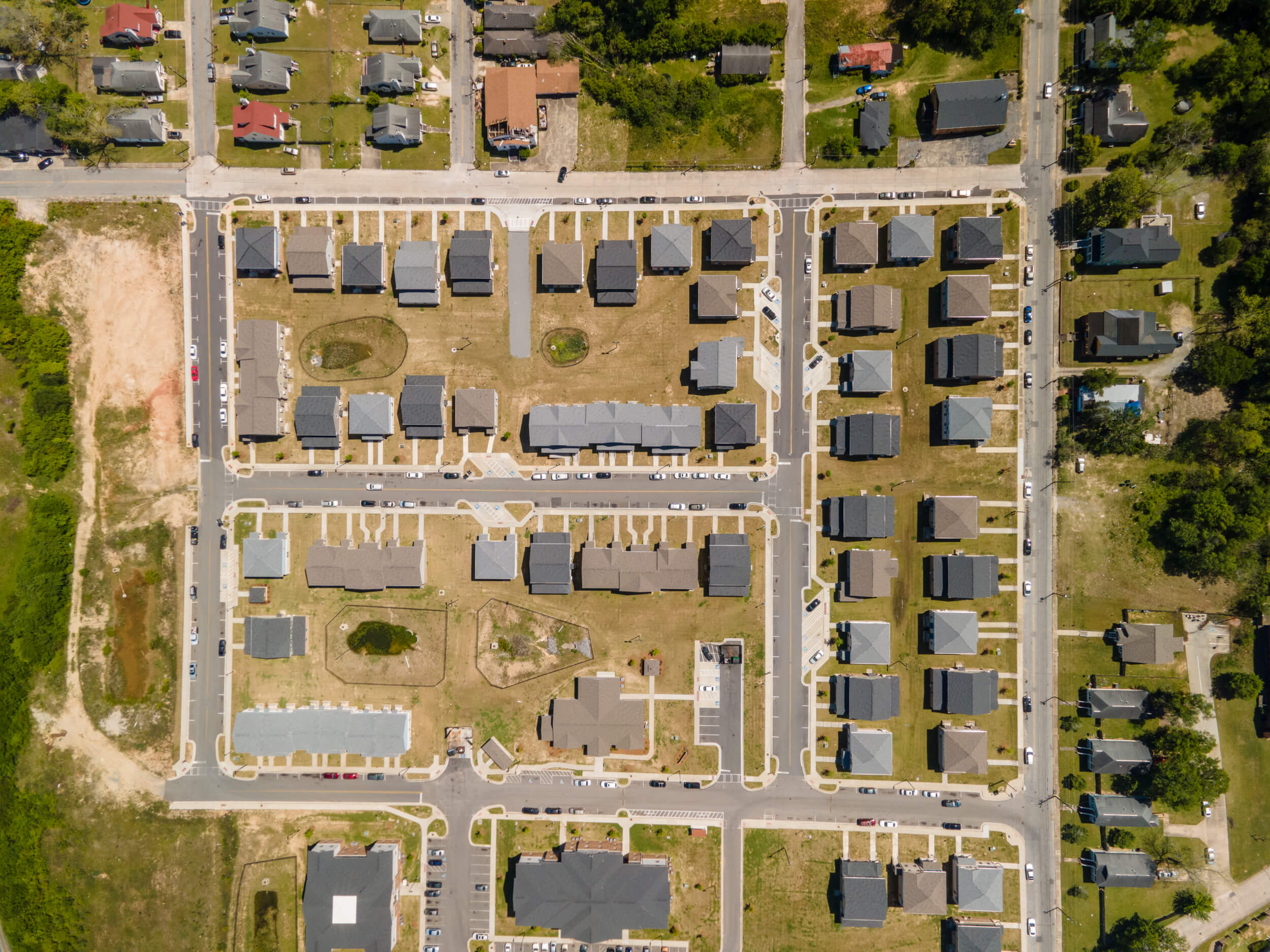 Aerial view of a suburban neighborhood with neat houses, streets, parked cars, grassy spaces, grid roads, and trees along the edge.