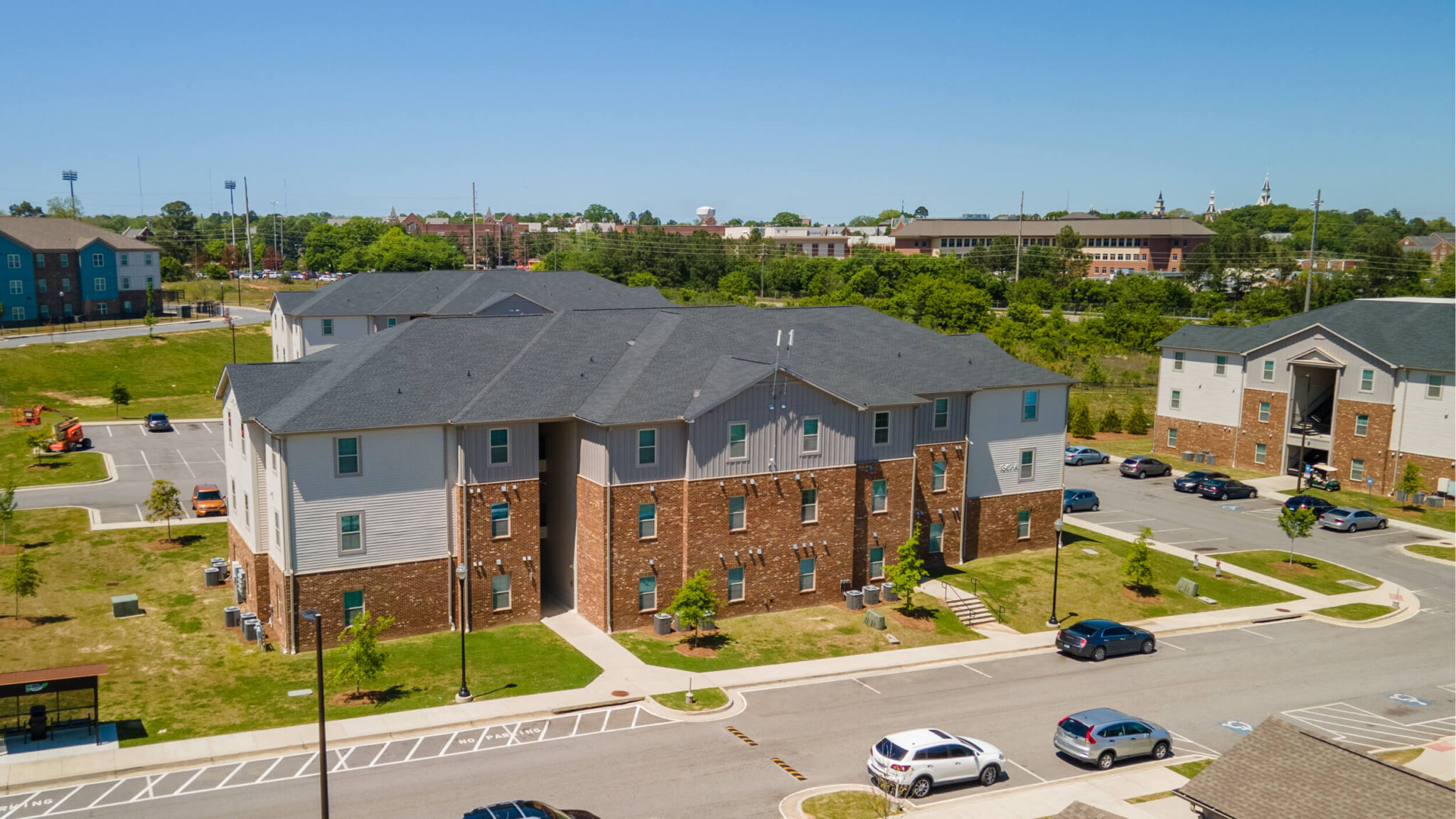 Aerial view of a modern brick and white apartment complex with parking, few cars, trees, and other buildings on a sunny day.
