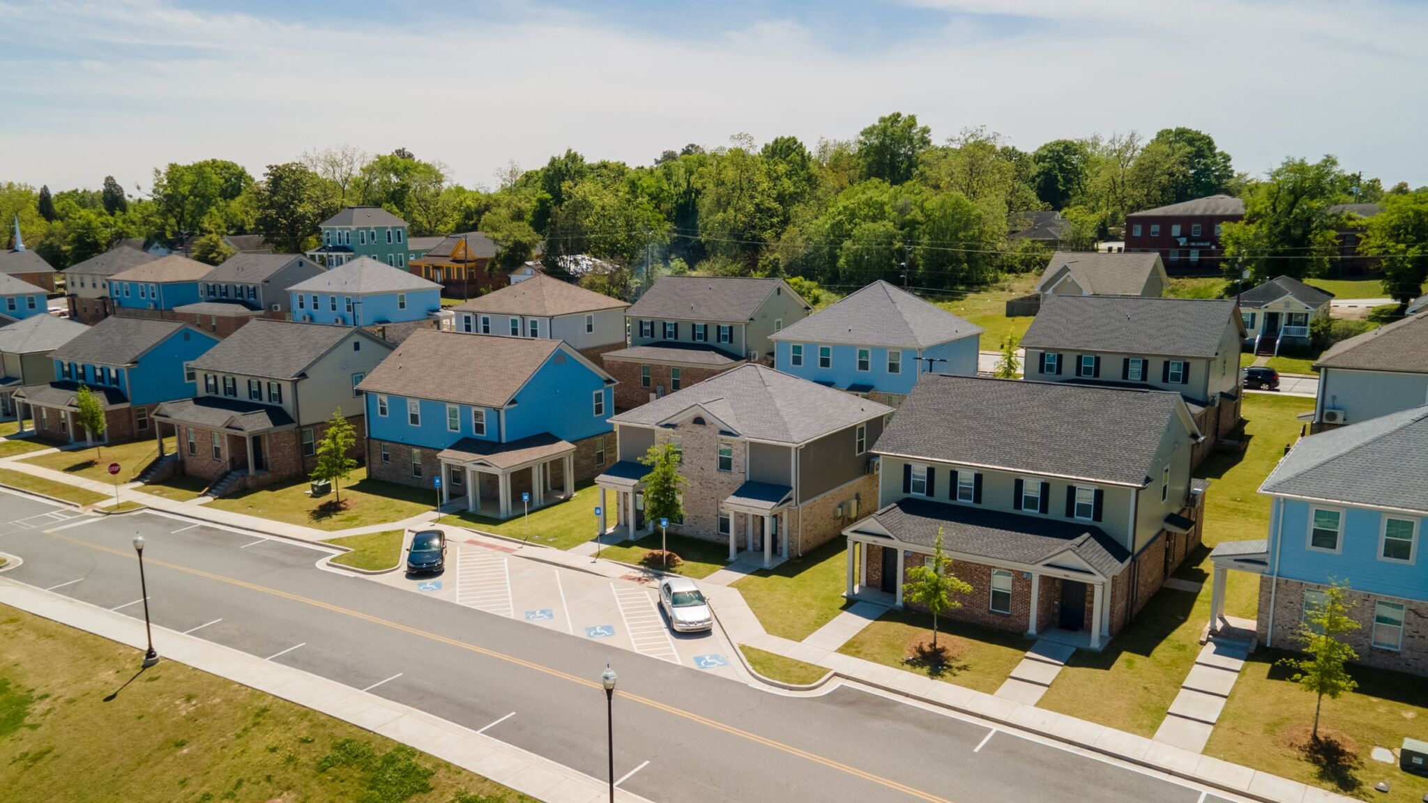 Row of modern suburban houses with driveways and lawns, viewed from above on a sunny day; parked cars and greenery in background.
