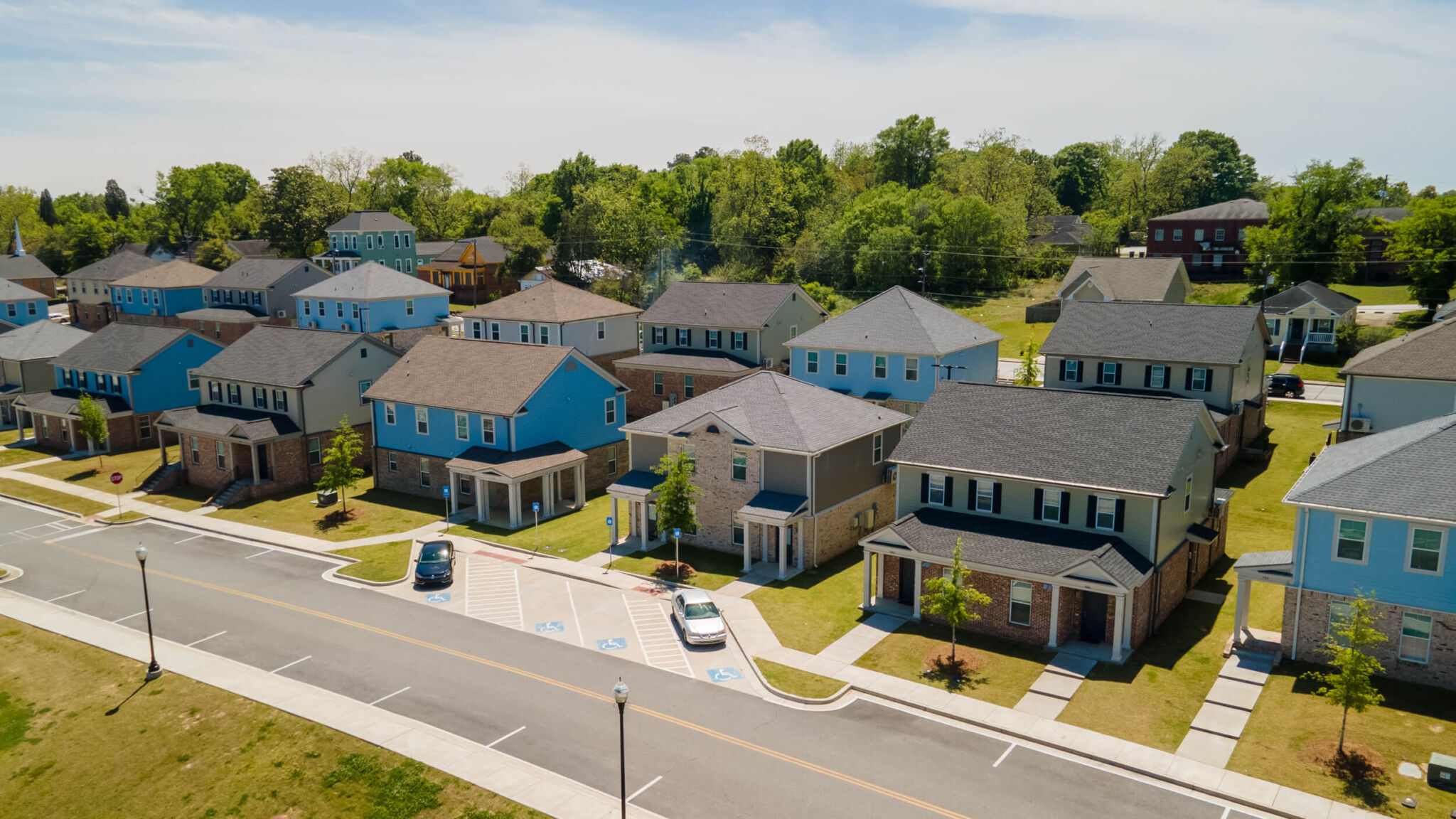 Aerial view of a suburban neighborhood, modern two-story houses, green lawns, parked cars on quiet streets under a clear sky.