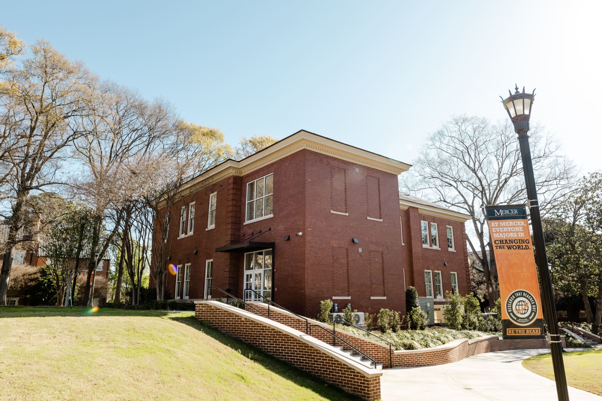 A two-story red brick building on a grassy lawn with trees, a ramp to the entrance, and a streetlamp with a Mercer University banner.
