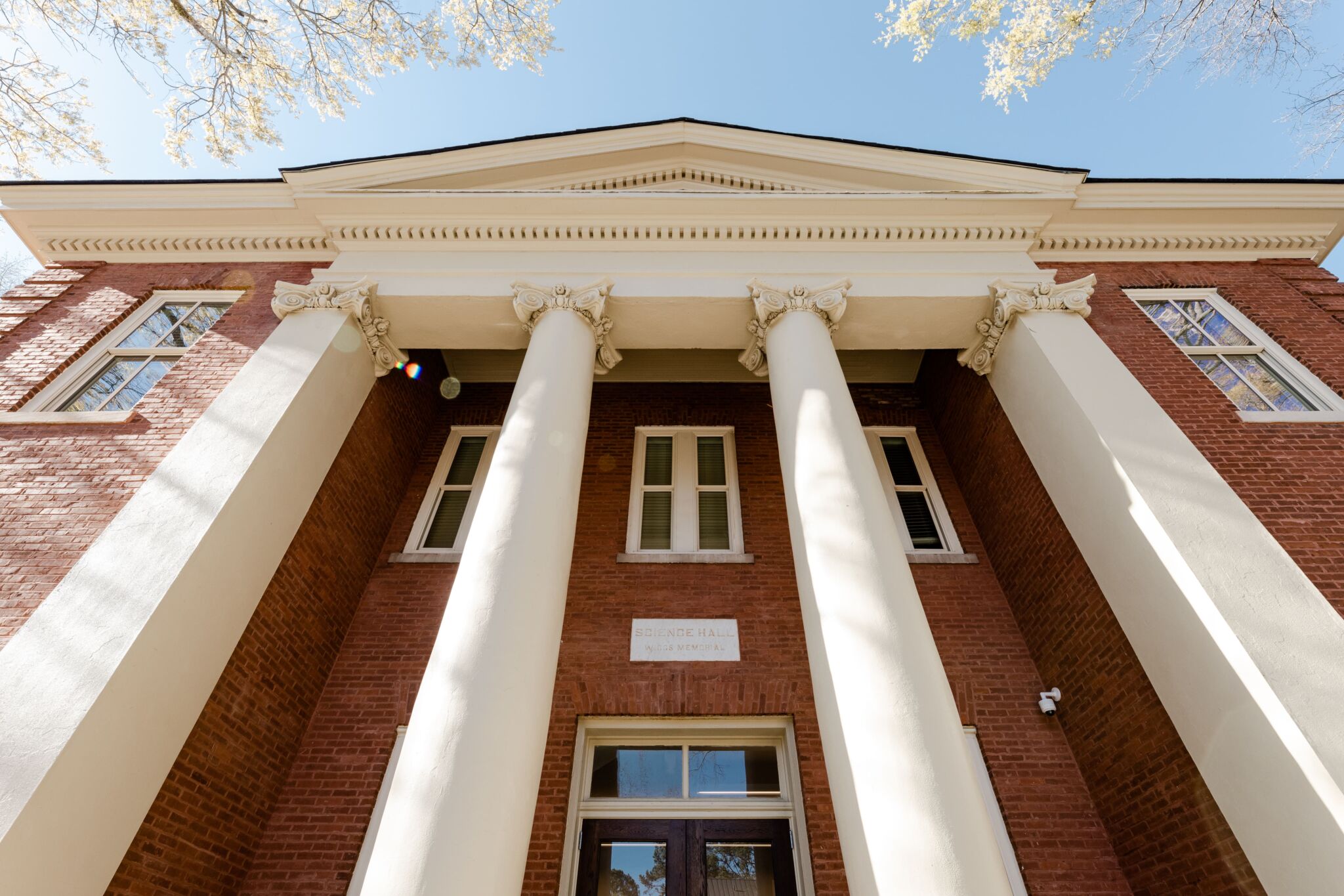 Low angle view of a red-brick building with white columns and tall windows under a blue sky, framed by tree branches at the top.