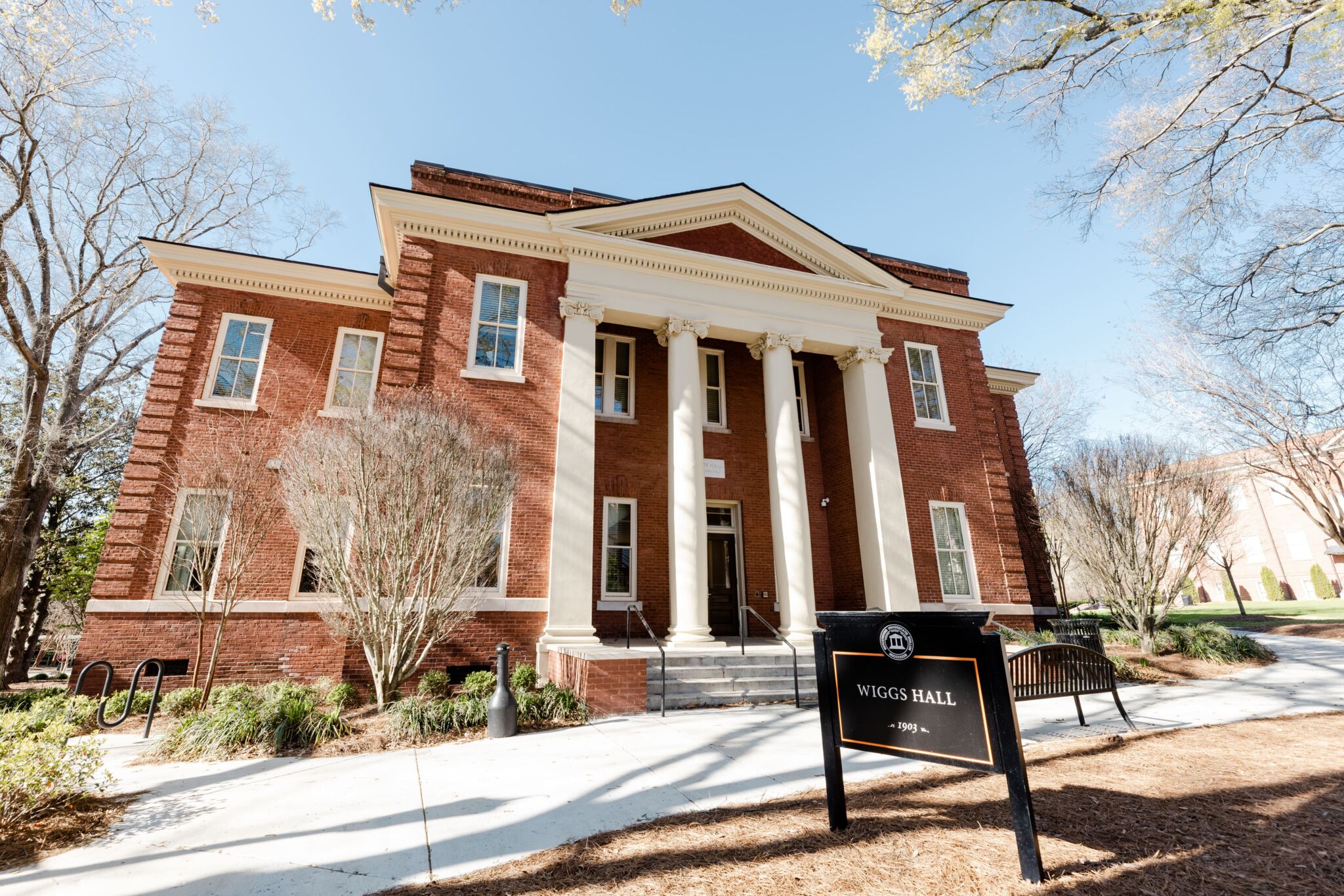 A red-brick building with large white columns at the entrance, labeled Wiggs Hall on a black sign. Trees and benches line the sidewalk on a sunny day.