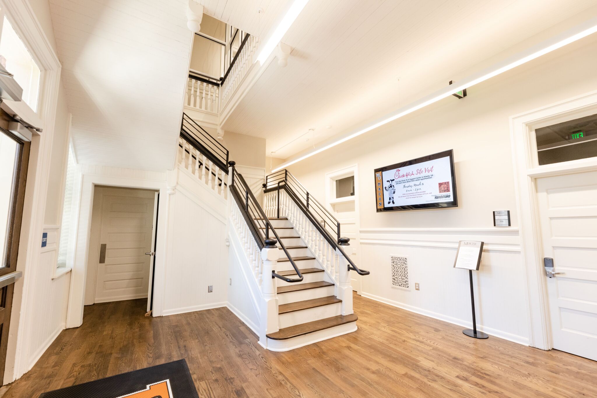 Bright, modern lobby with wooden floors, central staircase with black railings, white walls, doors, info screen, and large windows.