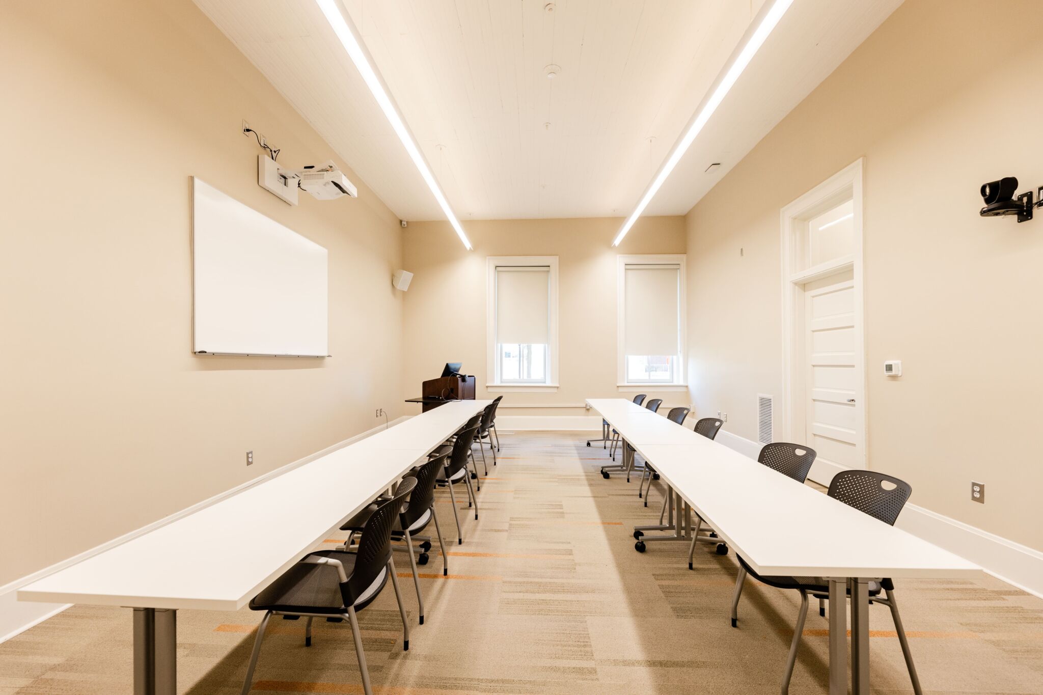 Modern classroom with beige walls, U-shaped white tables, black chairs, whiteboard, projector, ceiling lights, windows, podium with computer.