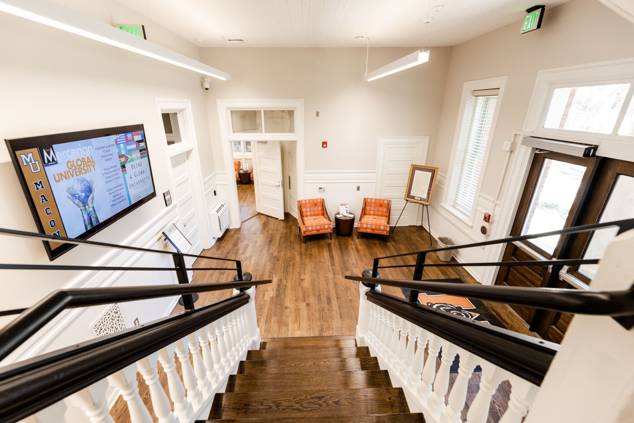 View from atop a staircase in a bright lobby with two orange chairs, an easel, large windows, entrance door, and left-wall display.
