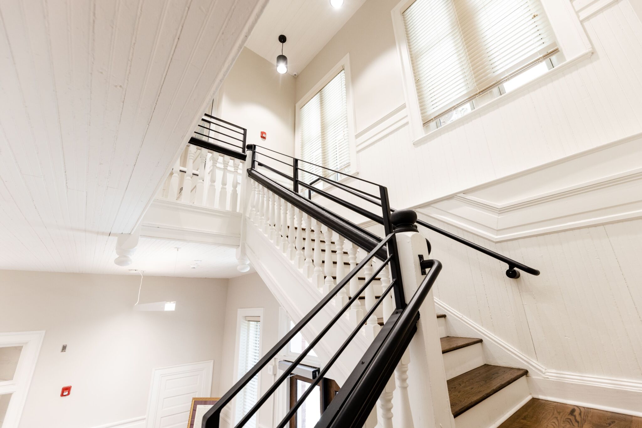 A bright modern staircase features white walls, wooden steps, black metal railings, and large windows letting in natural light.