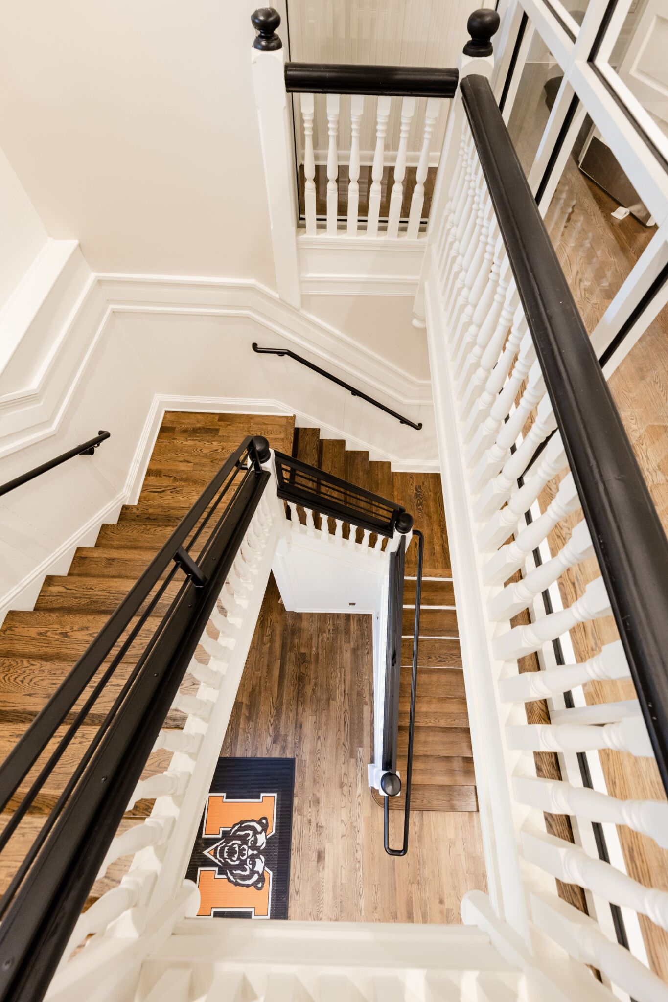 A view from above of a wooden staircase with white railings and black handrails, winding to a landing with a mat displaying M and an orange-black bear logo.