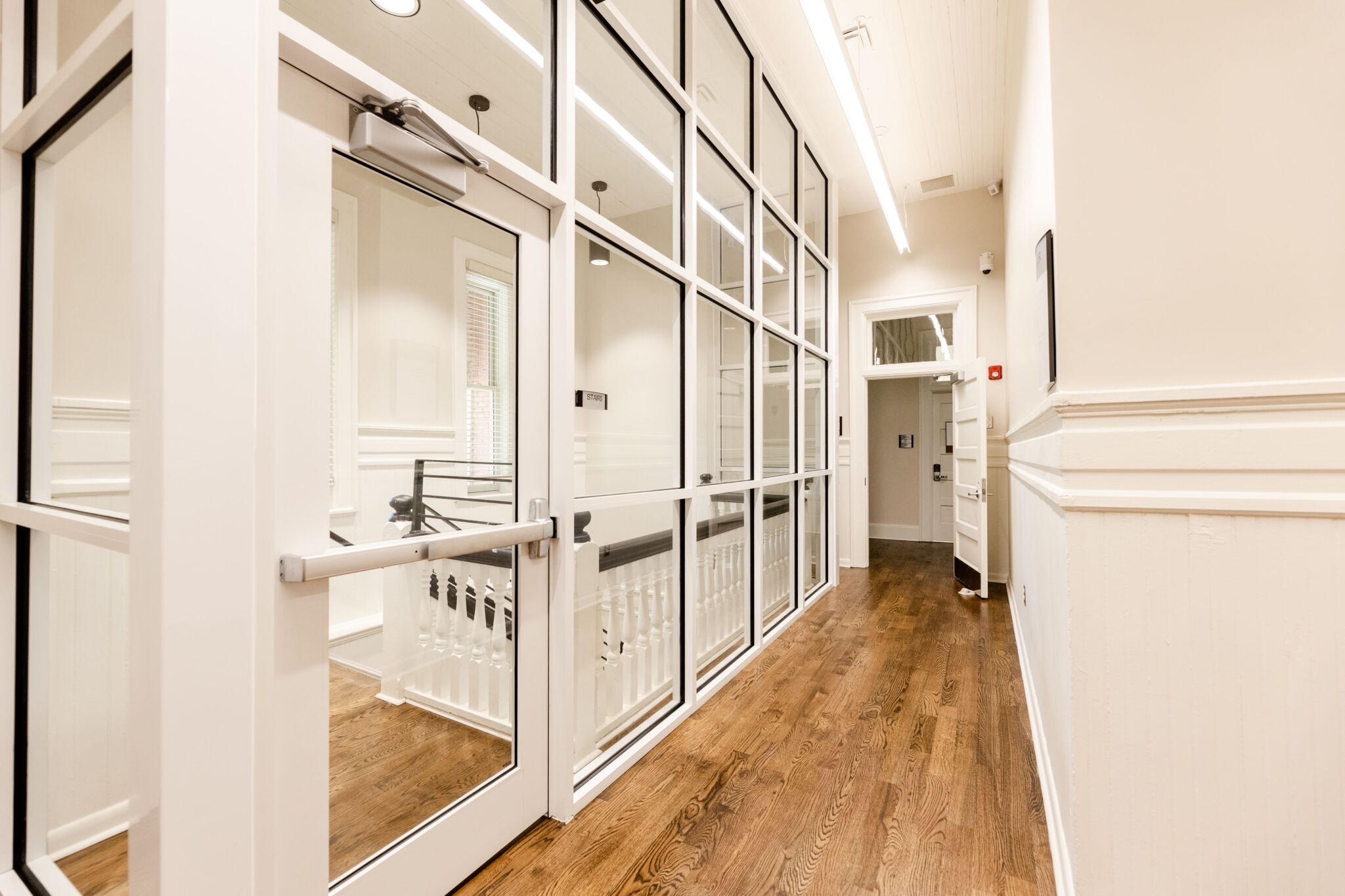 Clean, bright hallway with wood floors, white walls, and glass doors along one side. Sunlight streams in from the open doorway at the end.