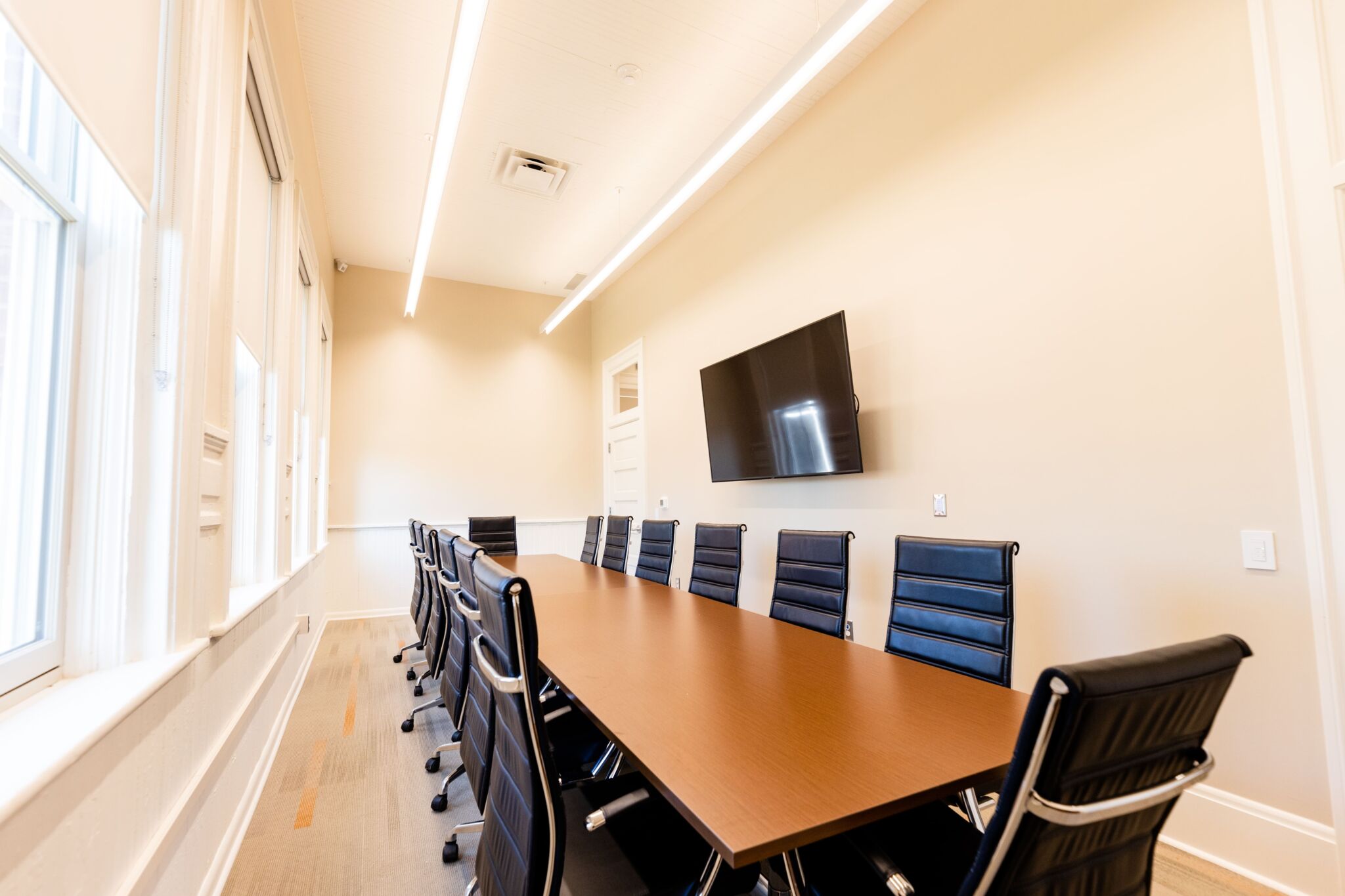 Modern conference room featuring a long wooden table, black office chairs, wall-mounted TV, large windows, and bright lighting.