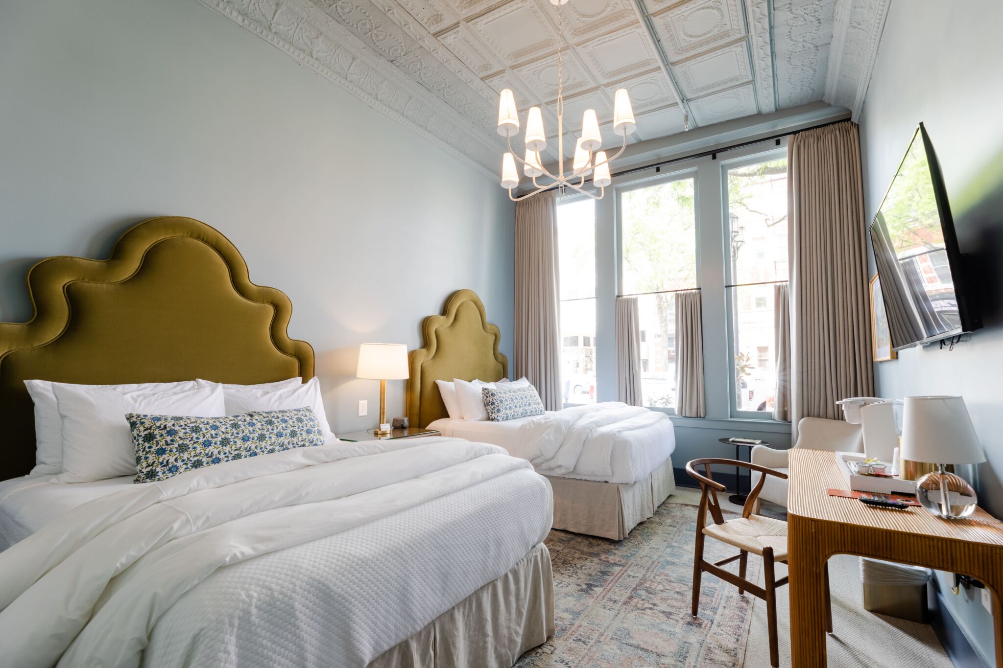Stylish hotel room with two beds, green ornate headboards, white bedding, chandelier, large windows, TV, desk, and lamp.