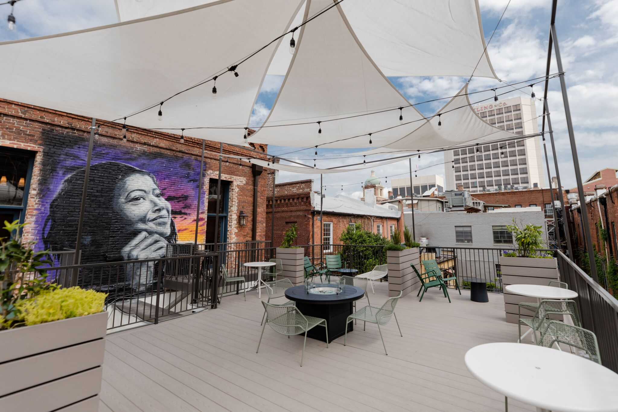 A rooftop patio with tables, chairs, string lights, a mural of a woman's face on brick, fabric shades above, and city buildings behind.