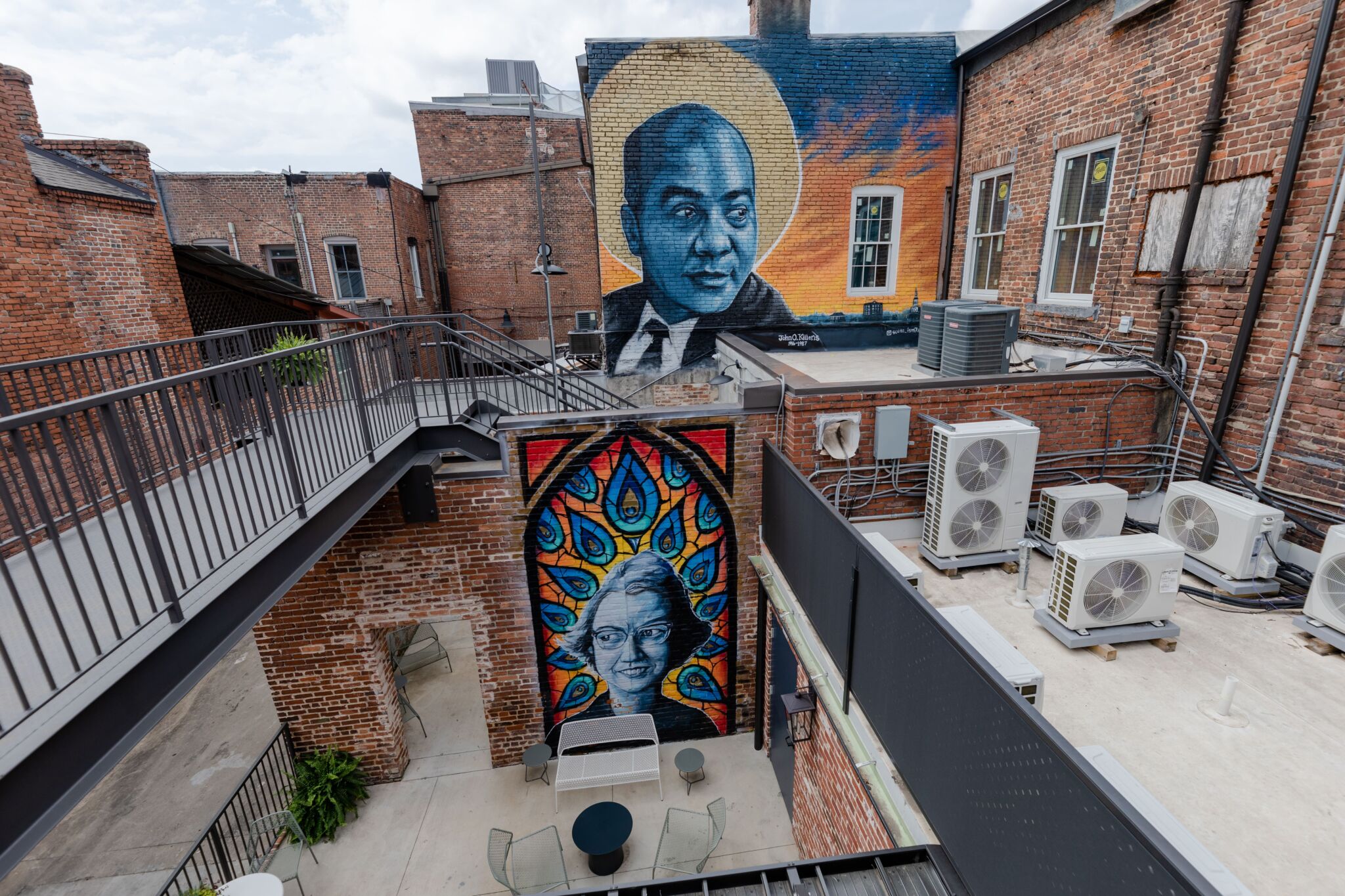 A brick courtyard displays two murals: a man with a gold halo above, and a woman with peacock feathers below. Metal stairs and seats.