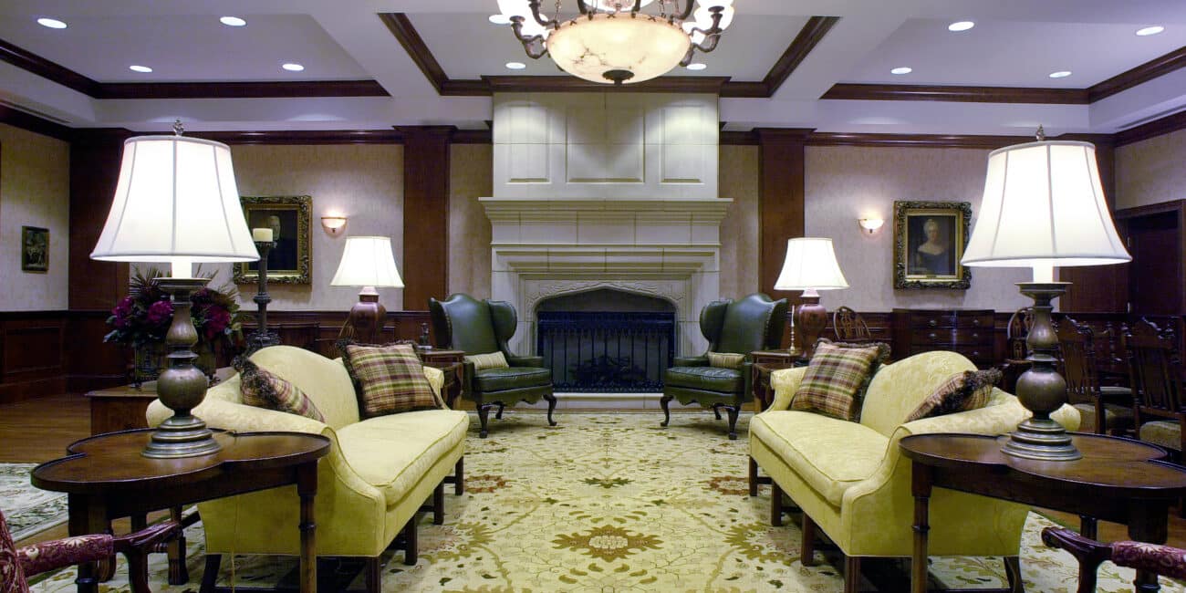 Elegant traditional lounge with yellow sofas, armchairs, tables, lamps, large fireplace, chandelier, wood paneling, and floral rug.