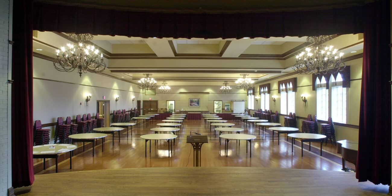 View from a stage showing a spacious, empty banquet hall with round tables, chandeliers overhead, and large windows on the right wall.