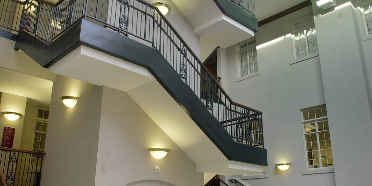 Bright modern atrium featuring several staircases, an elevator, skylights, and a round table with flowers on a patterned central rug.