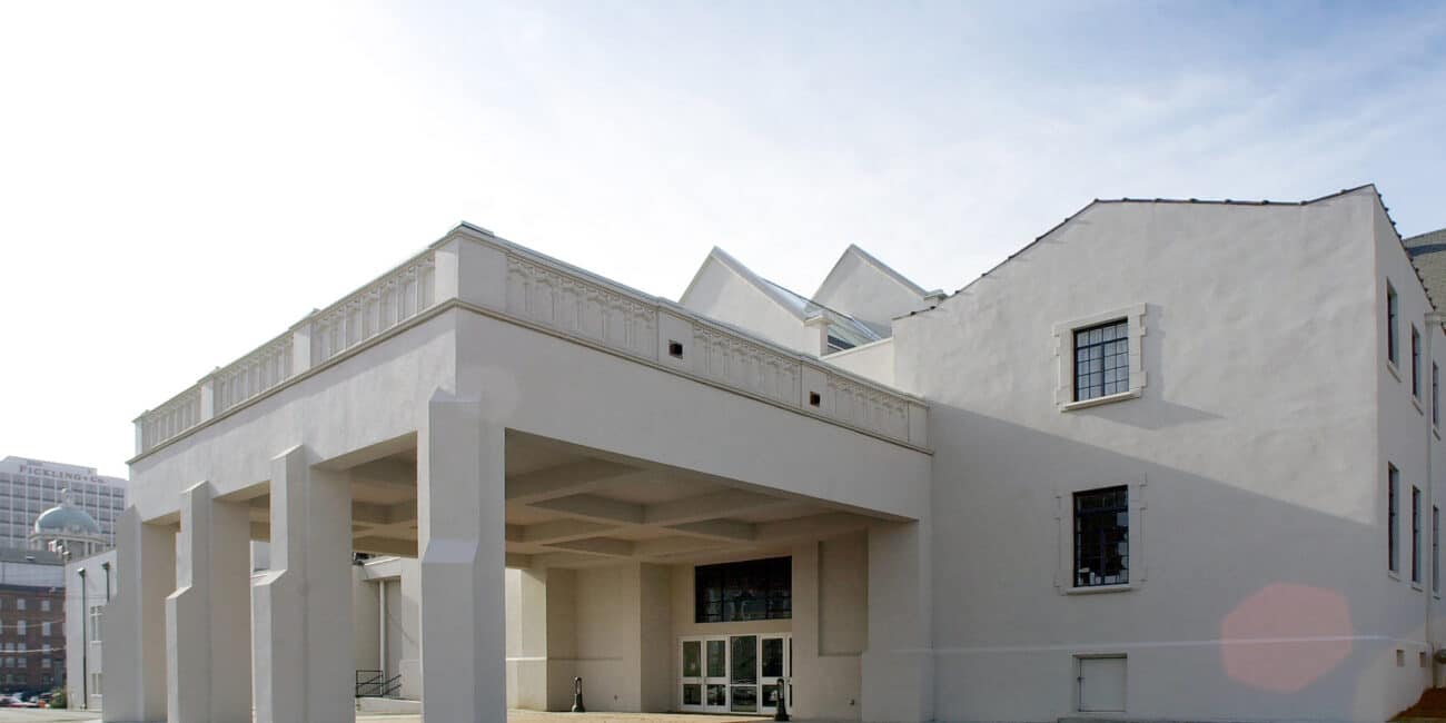 A white modern building with a pillar-supported entrance, flat geometric lines, and city buildings in the background under a clear sky.