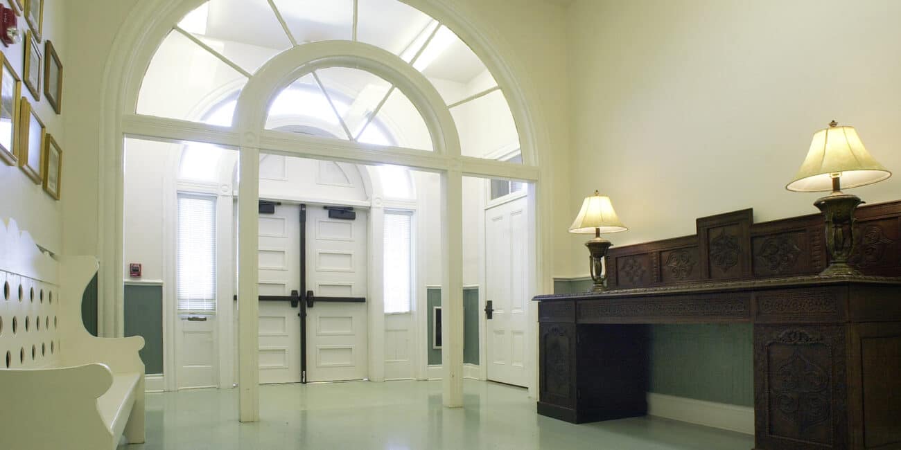 Bright hallway with high ceilings, double white doors with arched windows, a white bench, dark wood console and two lit lamps. Pale walls, light blue floor.