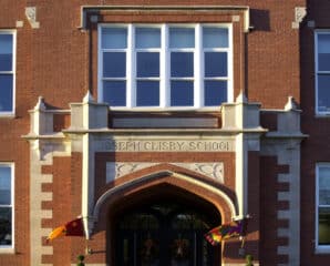 A red-brick school building with arched entrance, two flags, and Joseph Clisby School engraved above the doorway. Symmetrical windows and decorative stonework frame the entrance.