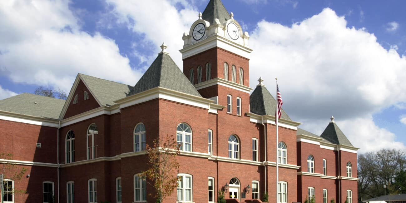 A large red brick courthouse with arched windows, a clock tower, gray roofs, American flag outside, cloudy sky, and nearby trees.