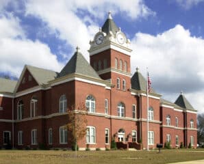 A large red brick courthouse with arched windows, a clock tower, gray roofs, American flag outside, cloudy sky, and nearby trees.