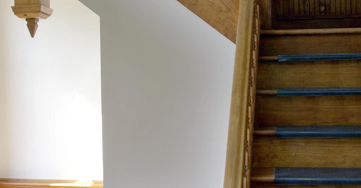 Wooden staircase with blue carpeted steps, wooden banister, matching wainscoting, all lit by natural light from a nearby window.