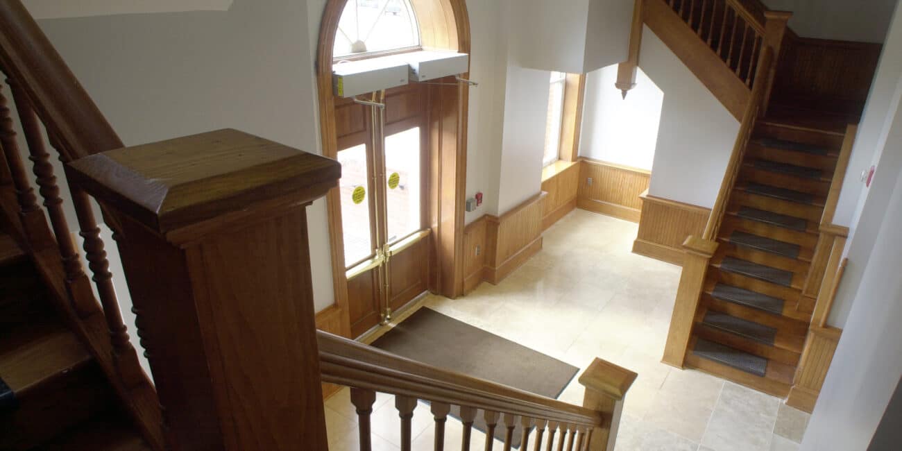Bright building lobby interior with wooden staircases, arched glass double doors, tiled floor, and sunlight from large windows.