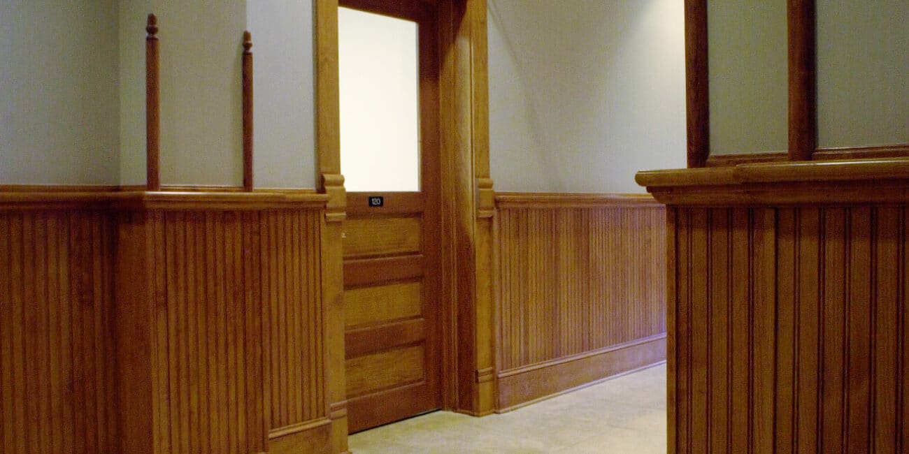 A hallway with wood-paneled walls and trim, beige tile floor, recessed ceiling lights, and a wooden door with frosted glass. A black directional sign hangs from the ceiling.