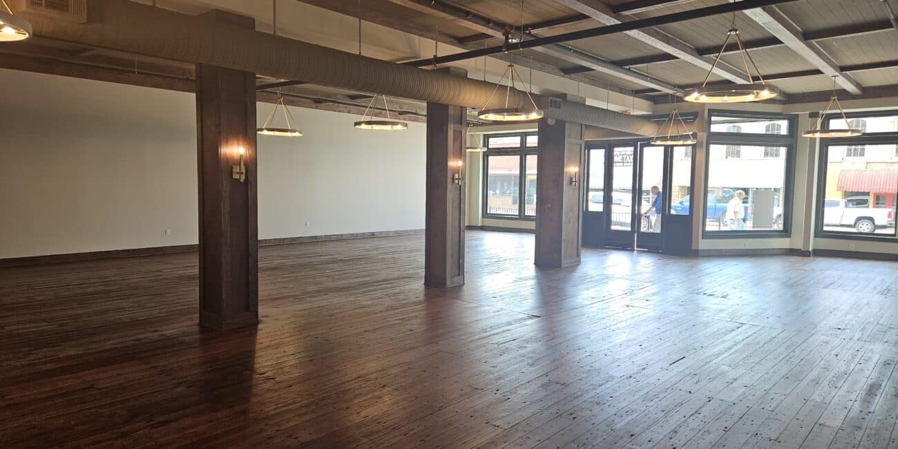 Spacious empty room with dark wood floors, exposed ceiling beams, large windows with natural light, and hanging lights. People outside.