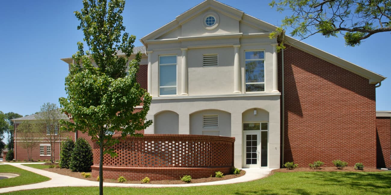 Modern two-story brick and stucco building with white trim and columns, set amid green grass, trees, landscaping, and blue sky.