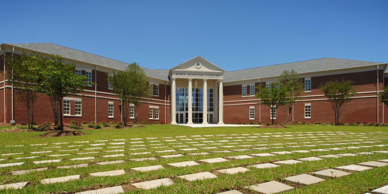 Red-brick two-story academic building with large columns and glass entrance, surrounded by grass, trees, walkway, under blue sky.